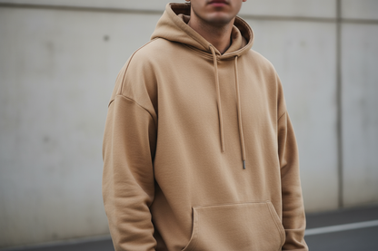 A minimalist streetwear hoodie in warm beige color, 450gsm heavyweight cotton, relaxed fit with dropped shoulders and visible drawstrings on the hood, photographed outdoors in soft daylight, clean background, premium lifestyle product photo.